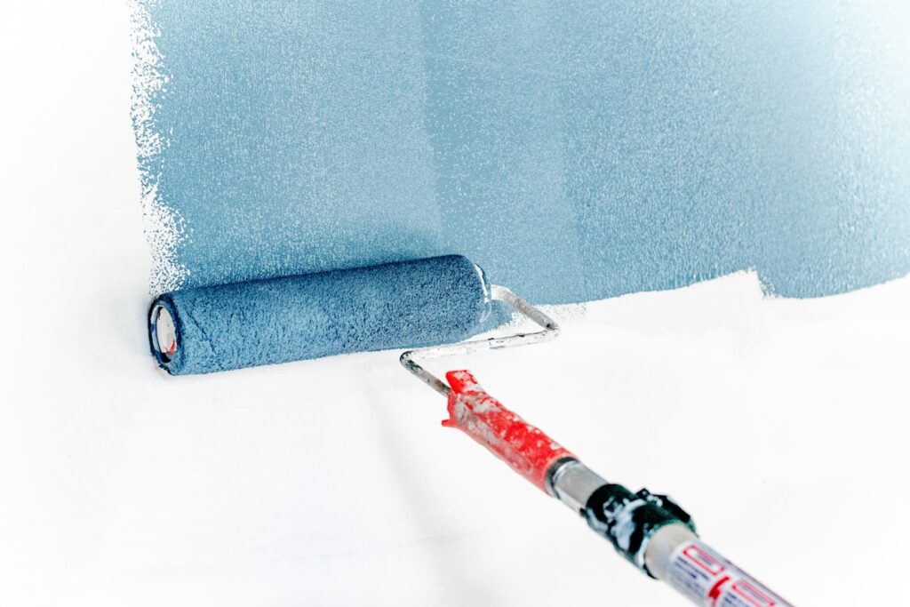 Blue paint being rolled onto an empty white wall with an extended paint roller